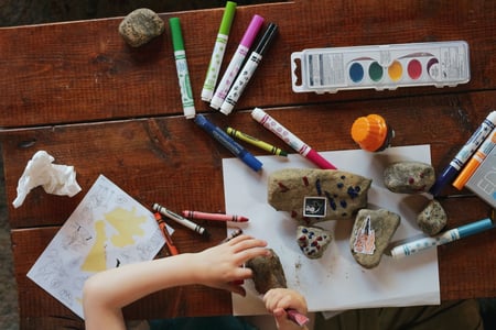 Photo looks down from above onto a table that has crayons, apper, markers, paints, rocks, and a set of hands trying to break open the rocks.