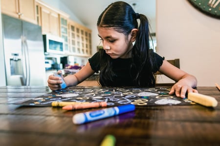 A young girl with black hair in pigtails sits at a table, drawing on paper with chalk, with a marker and crayons in the foreground.