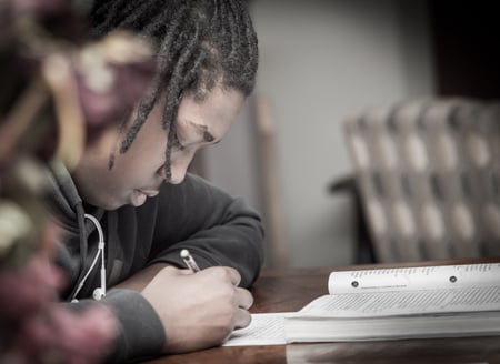 A young man of color sits at a table, his earbuds sticking out of his sweatshirt, while he writes, and a book is open on the table in front of him
