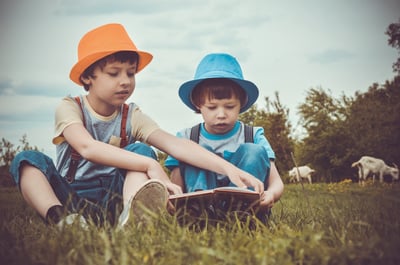 two boys wearing orange and blue hats while reading in a meadow