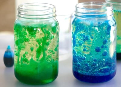 Two glass containers each contain a mixture of clear liquid and a colored liquid. The jar on the left has green "lava" and the jar on the right has blue.