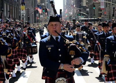 A man plays bagpipes while marching in a St. Patrick's Day parade. He is dressed in an Irish kilt and dress shirt and blazer up top.